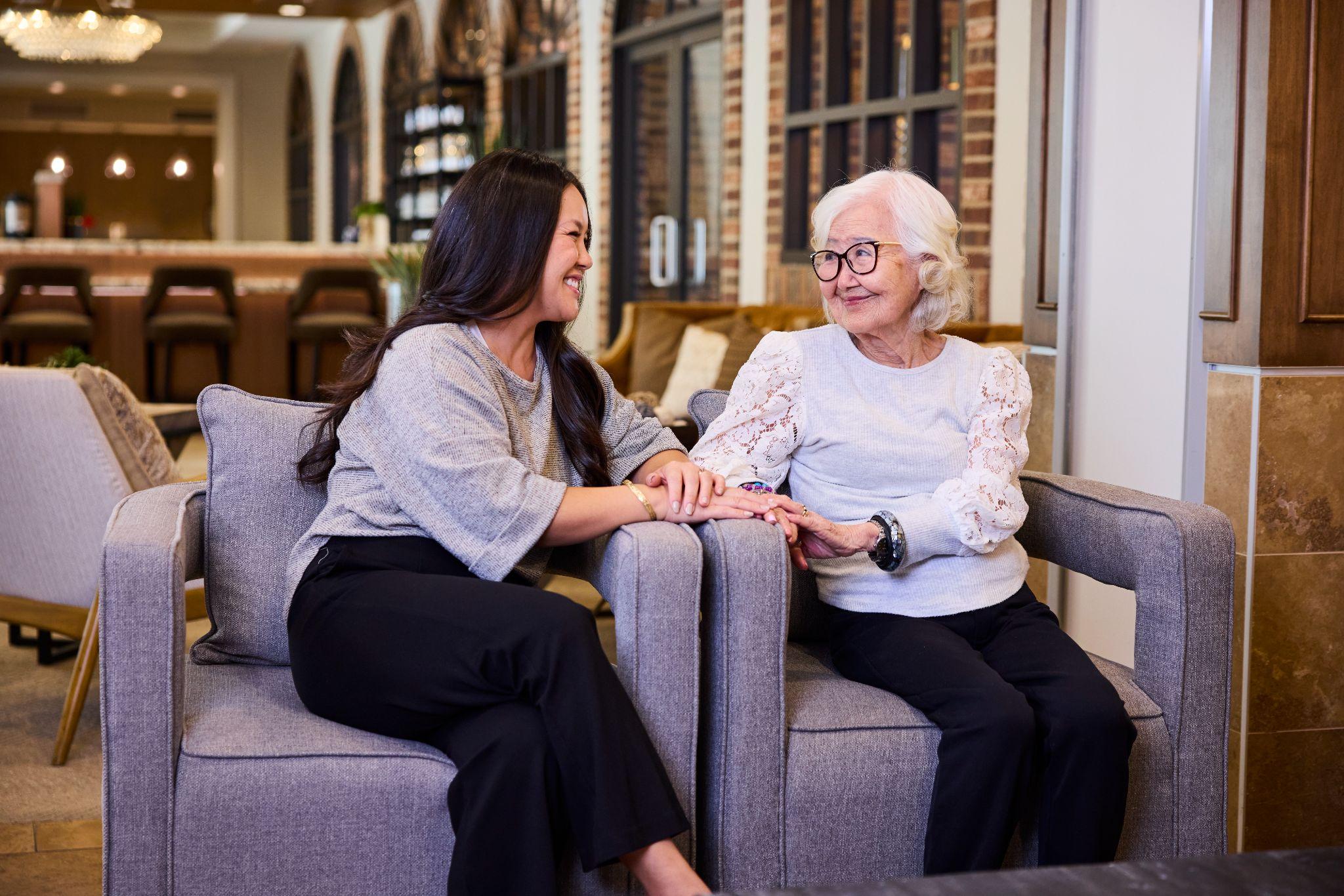 Older adult and senior parent sitting together, hand in hand while looking at each other, on two couches in an independent senior living community